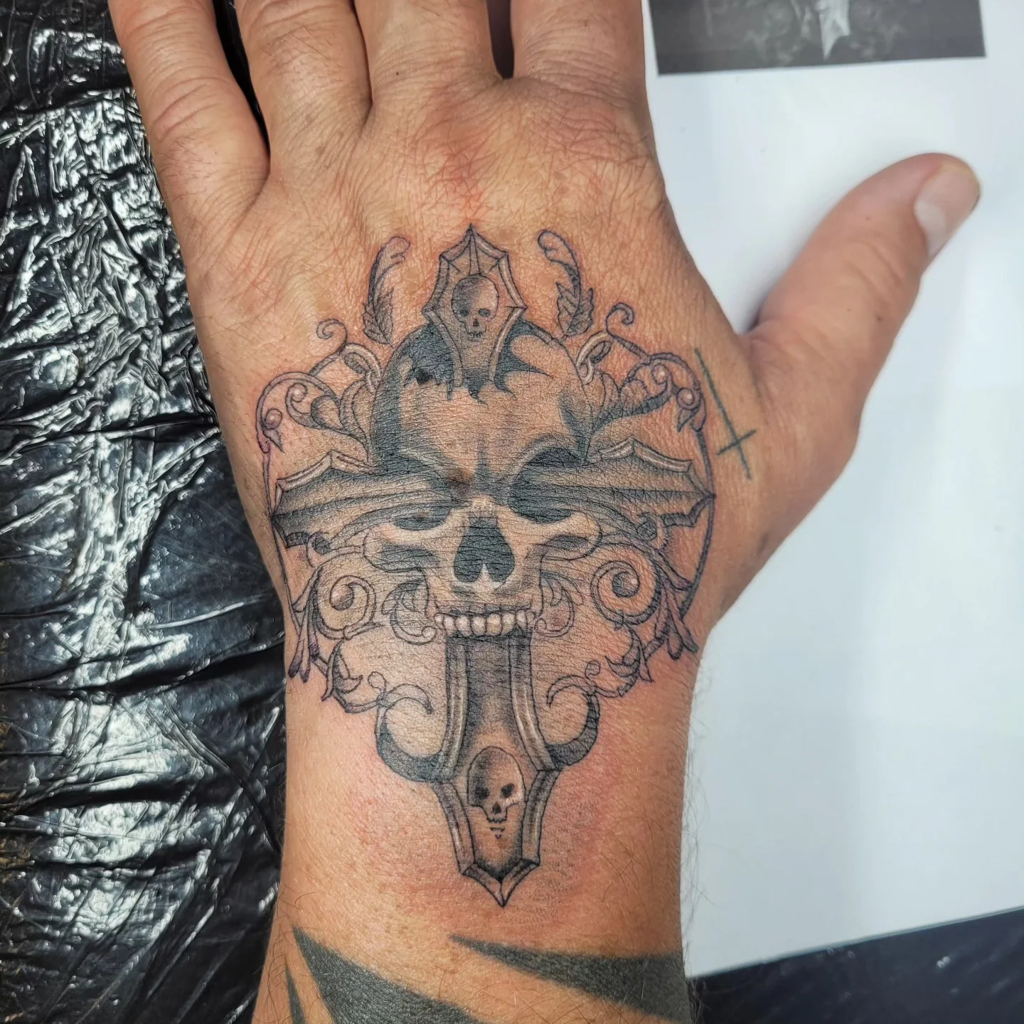 Tatouage chicano d'un crâne orné sur une croix gothique, sur le dos de la main, noir et gris, réalisé par Jean-Marie Joullié au salon JMJ Tattoo à Béziers et Sérignan