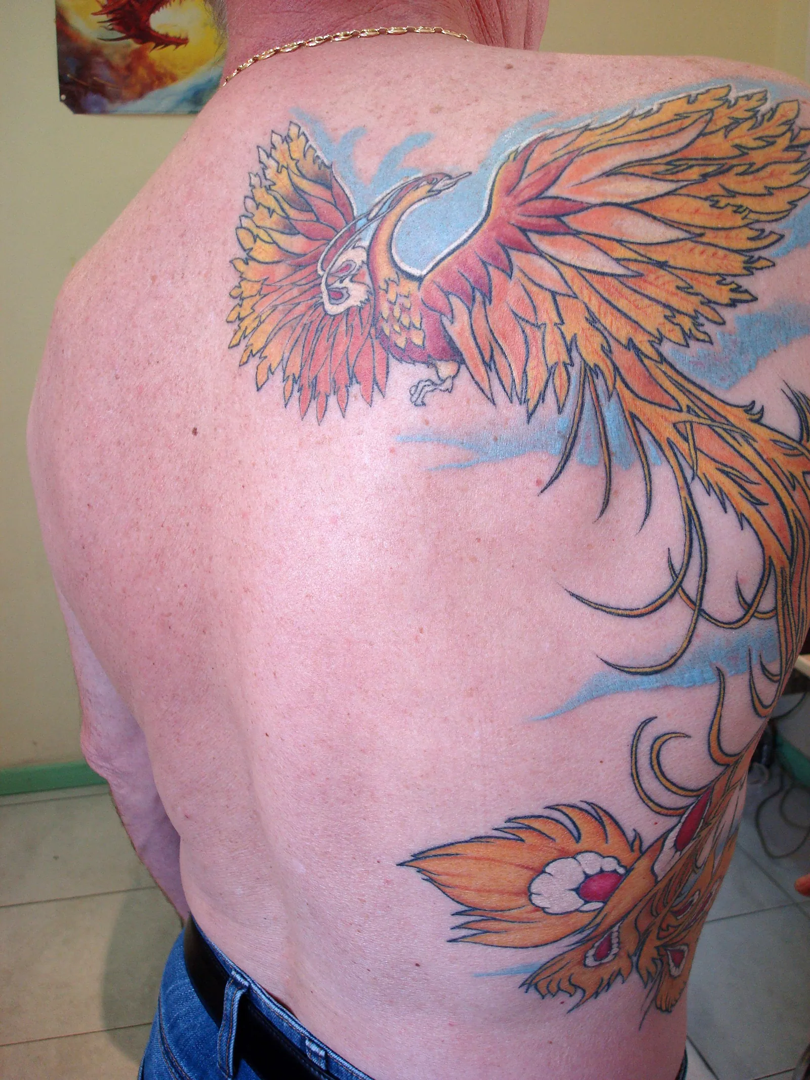 Tatouage couleur grand format d'un phénix flamboyant orange et jaune aux ailes déployées sur le haut du dos, queue qui retombe en plumes ardentes, réalisé par Jean-Marie Joullié au salon JMJ Tattoo à Sérignan