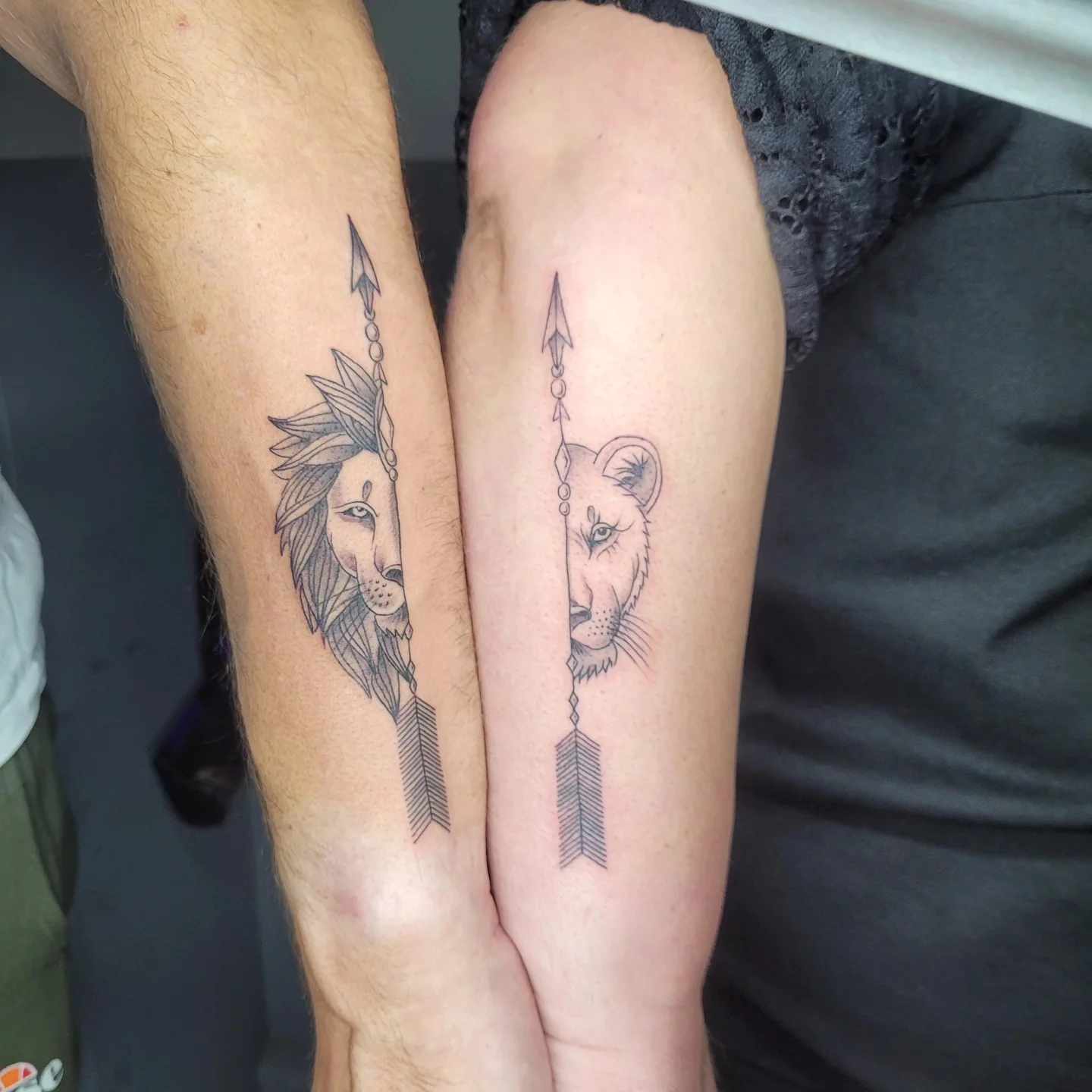 Tatouages fine line assortis sur deux avant-bras : lion et lionne stylisés avec flèche géométrique, réalisés par Jean-Marie Joullié au salon JMJ Tattoo proche de Cers