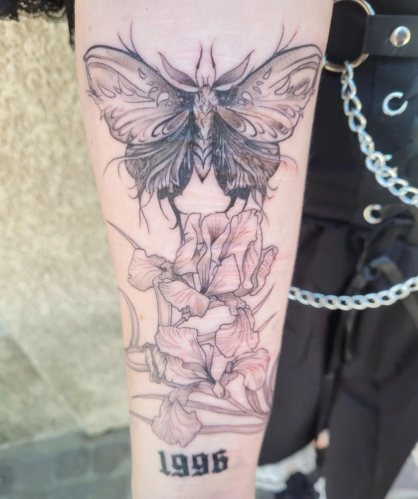 Tatouage fine line d'un papillon de nuit au-dessus d'iris, avec date « 1996 » en lettrage gothique, sur l'avant-bras, réalisé par Jean-Marie Joullié au salon JMJ Tattoo proche de Cap d'Agde