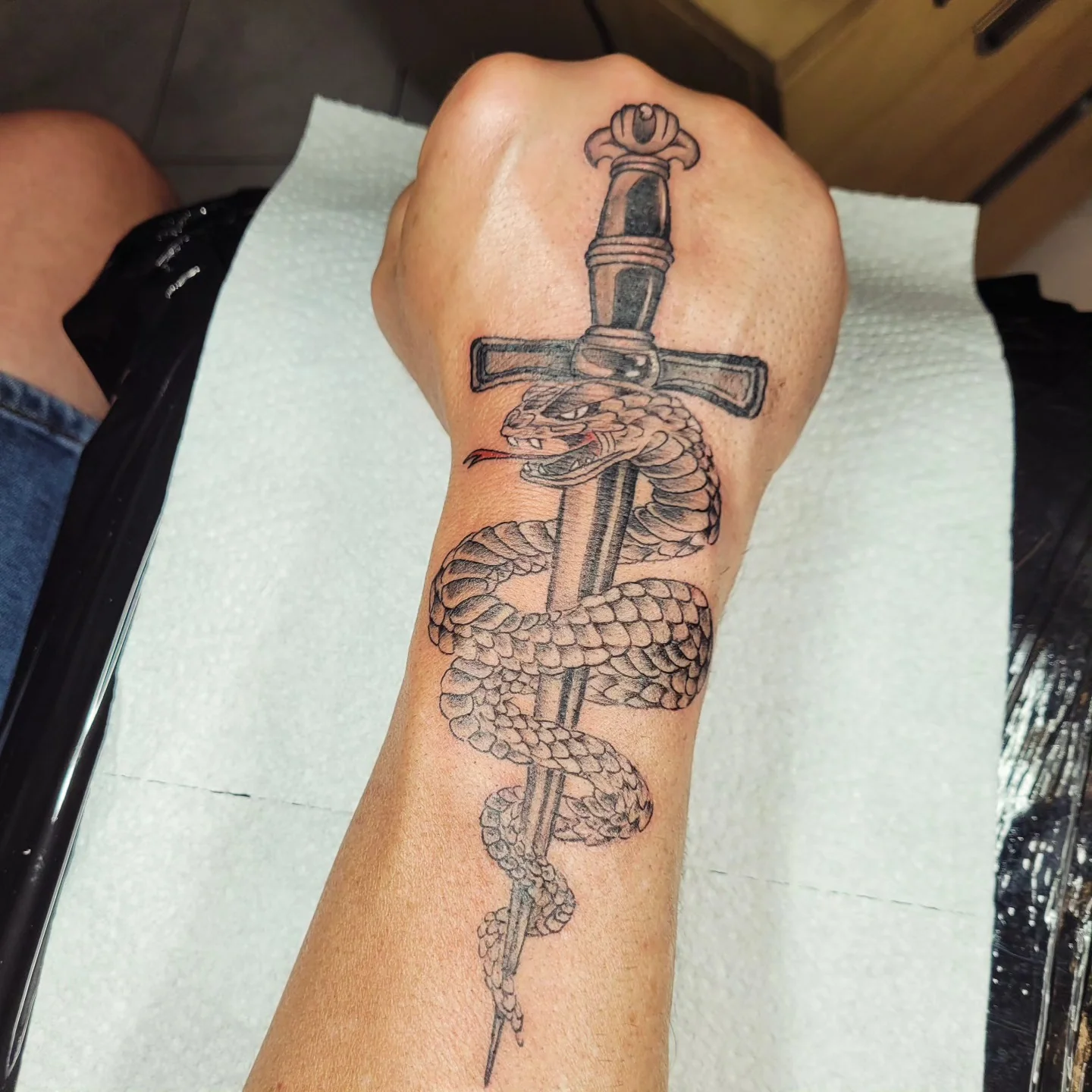 Tatouage d'un serpent enroulé autour d'une épée avec garde en forme de croix, poignet et main en noir et gris inspiration japonaise, réalisé par Jean-Marie Joullié au salon JMJ Tattoo près de Villeneuve-lès-Béziers