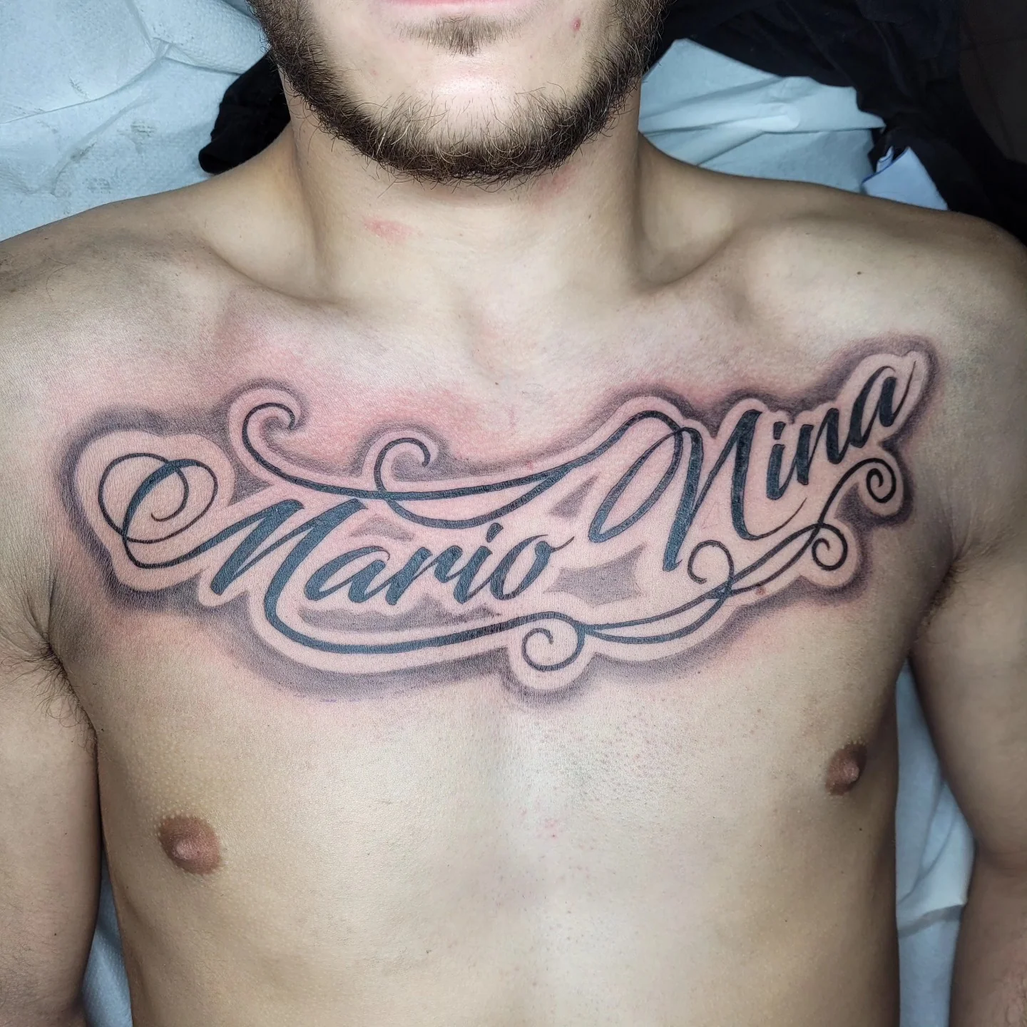 Tatouage de lettrage script sur la poitrine avec les prénoms « Mario Nina » en calligraphie élégante, ombres noir et gris, réalisé par Jean-Marie Joullié au salon JMJ Tattoo à Sérignan
