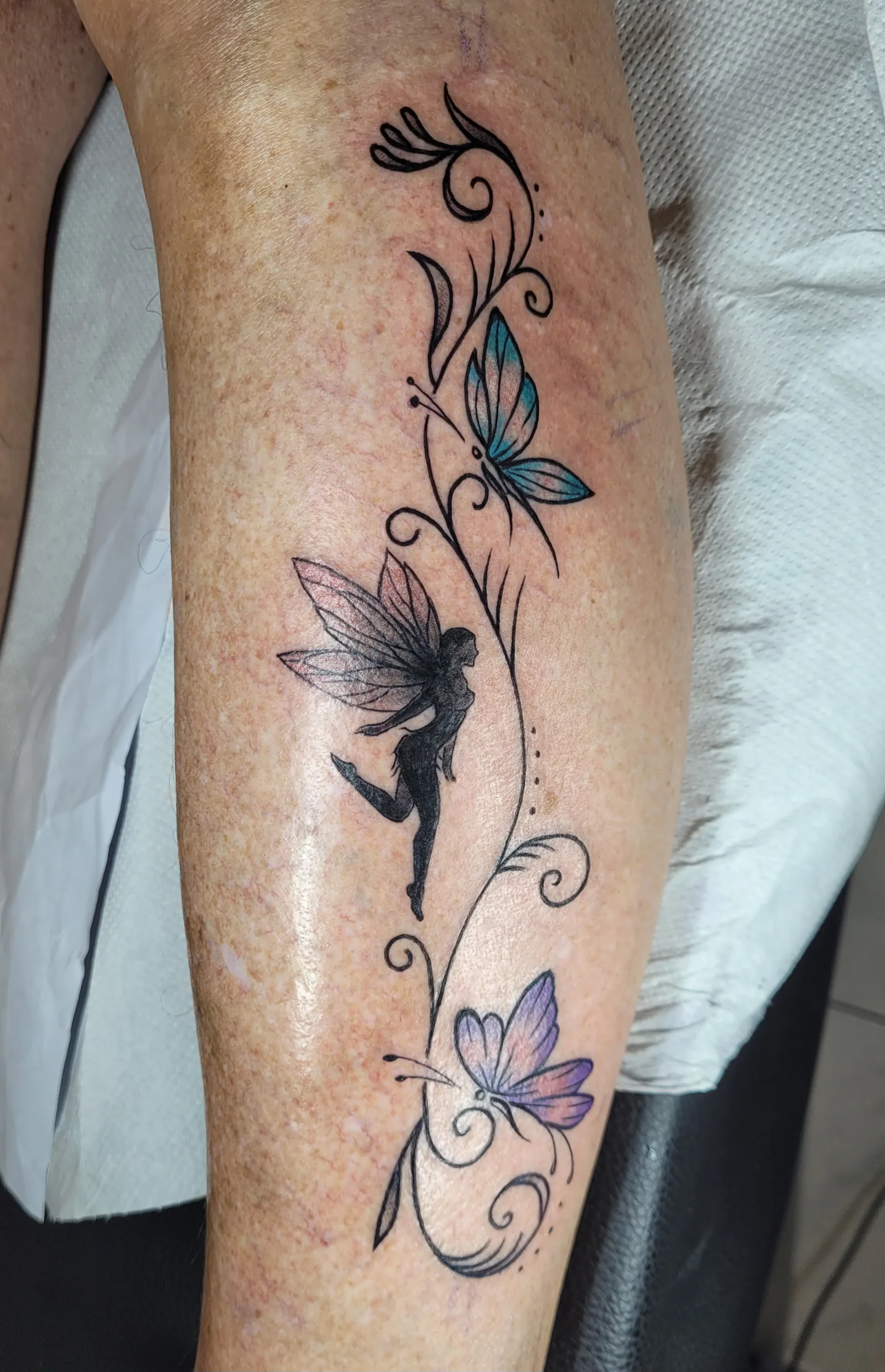 Tatouage noir et gris d'une fée entourée de papillons sur le mollet, réalisé par Jean-Marie Joullié au salon JMJ Tattoo proche de Portiragnes