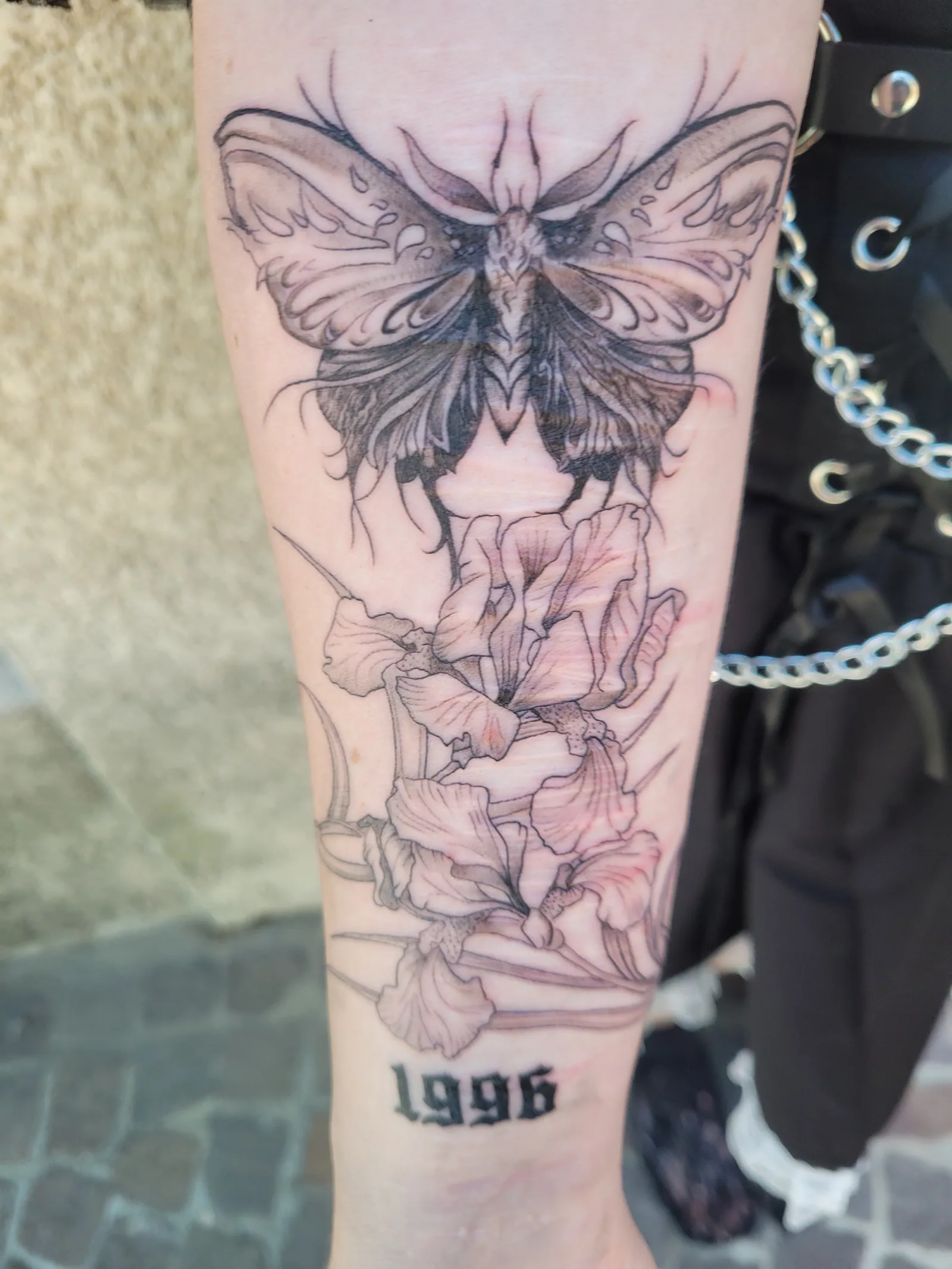 Tatouage noir et gris d'un papillon, d'iris et de la date 1996 sur l'avant-bras, pièce commémorative, réalisé par Jean-Marie Joullié au salon JMJ Tattoo proche de Pézenas