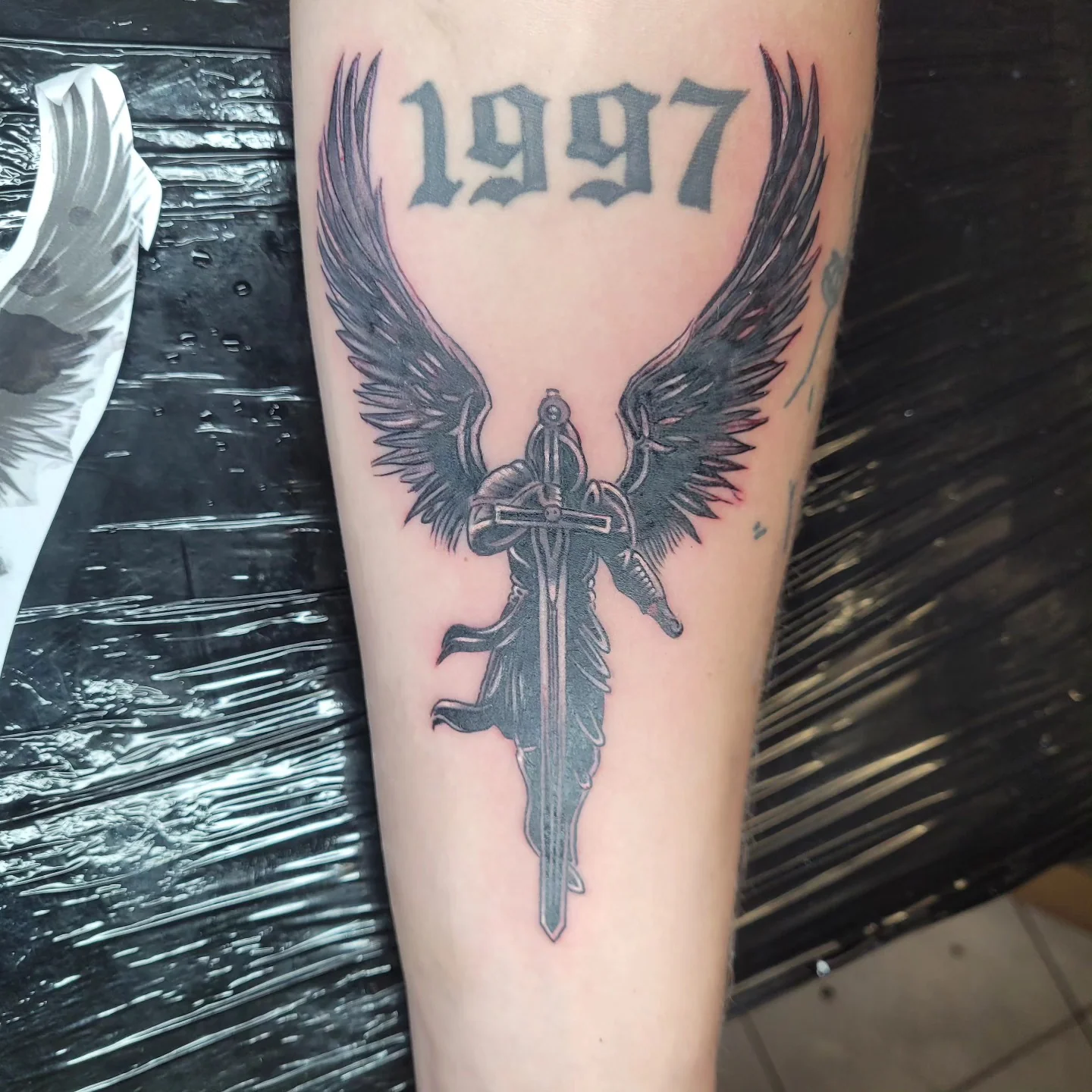 Tatouage d'un ange guerrier tenant une épée, ailes déployées, surmonté du lettrage gothique « 1997 » en hommage à la date d'ouverture du salon, avant-bras noir et gris, réalisé par Jean-Marie Joullié au salon JMJ Tattoo proche de Cers