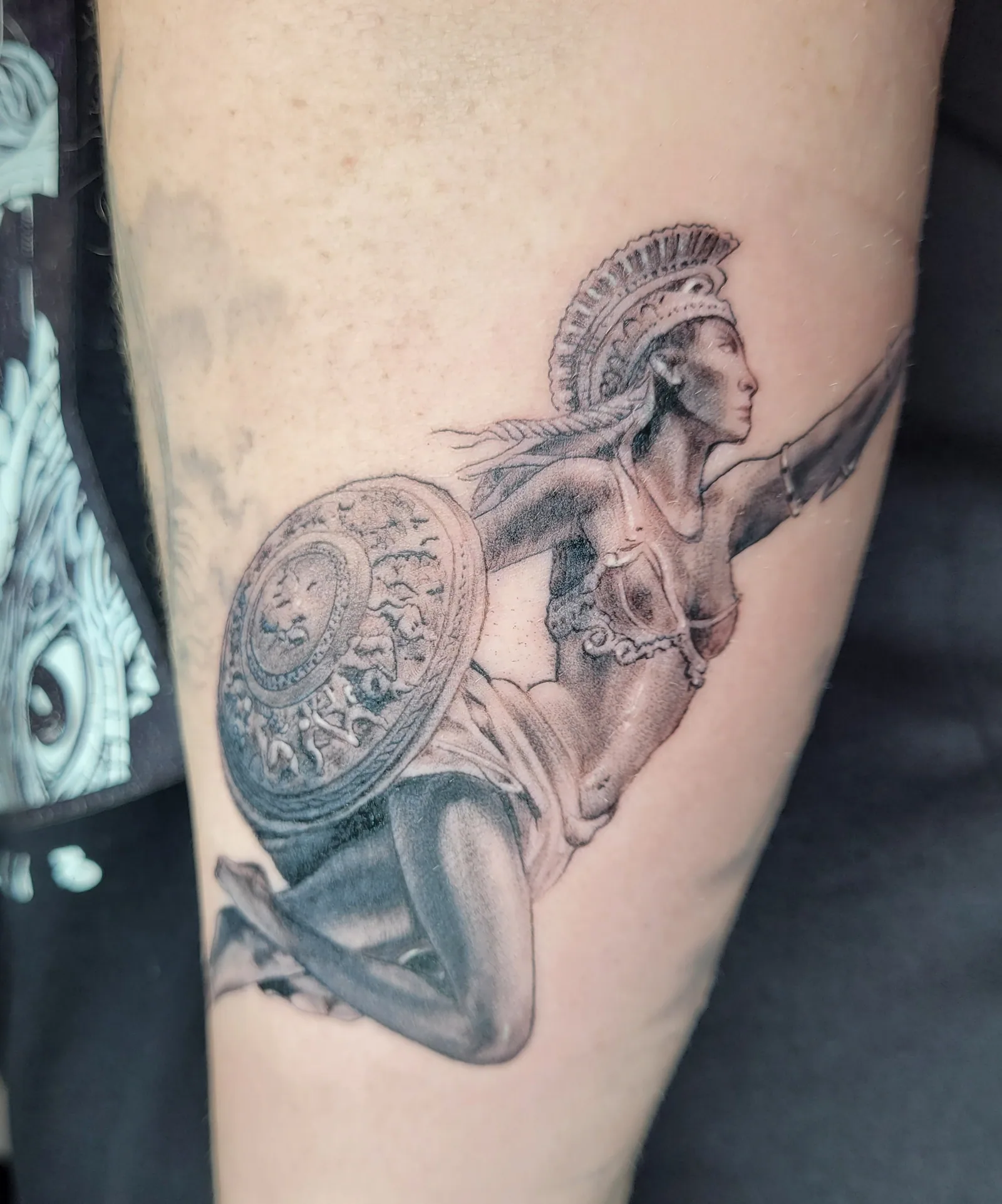 Tatouage réaliste d'une guerrière antique casquée avec bouclier sur la cuisse, en noir et gris, réalisé par Jean-Marie Joullié au salon JMJ Tattoo proche d'Agde