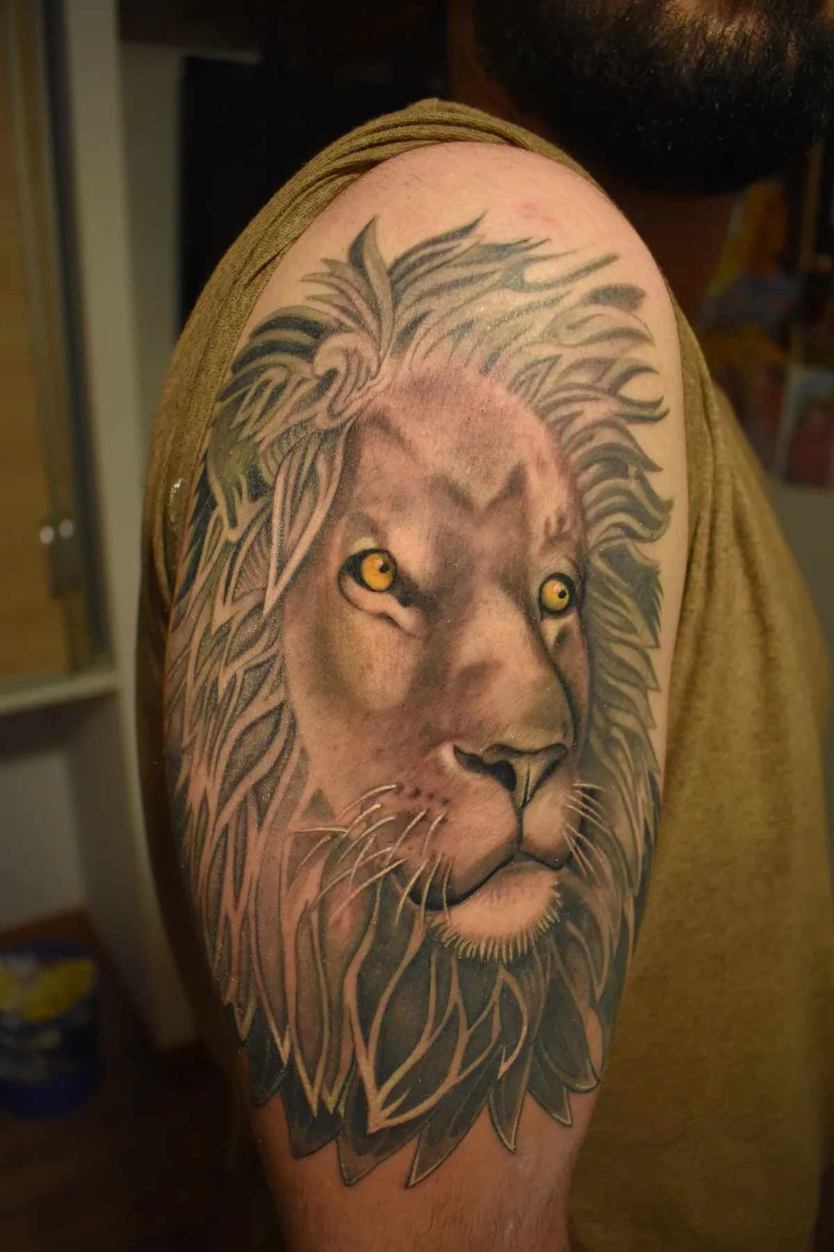 Tatouage réaliste d'une tête de lion noir et gris au regard doré, sur l'épaule, crinière flamboyante et expression apaisée, réalisé par Jean-Marie Joullié au salon JMJ Tattoo à Béziers et Sérignan