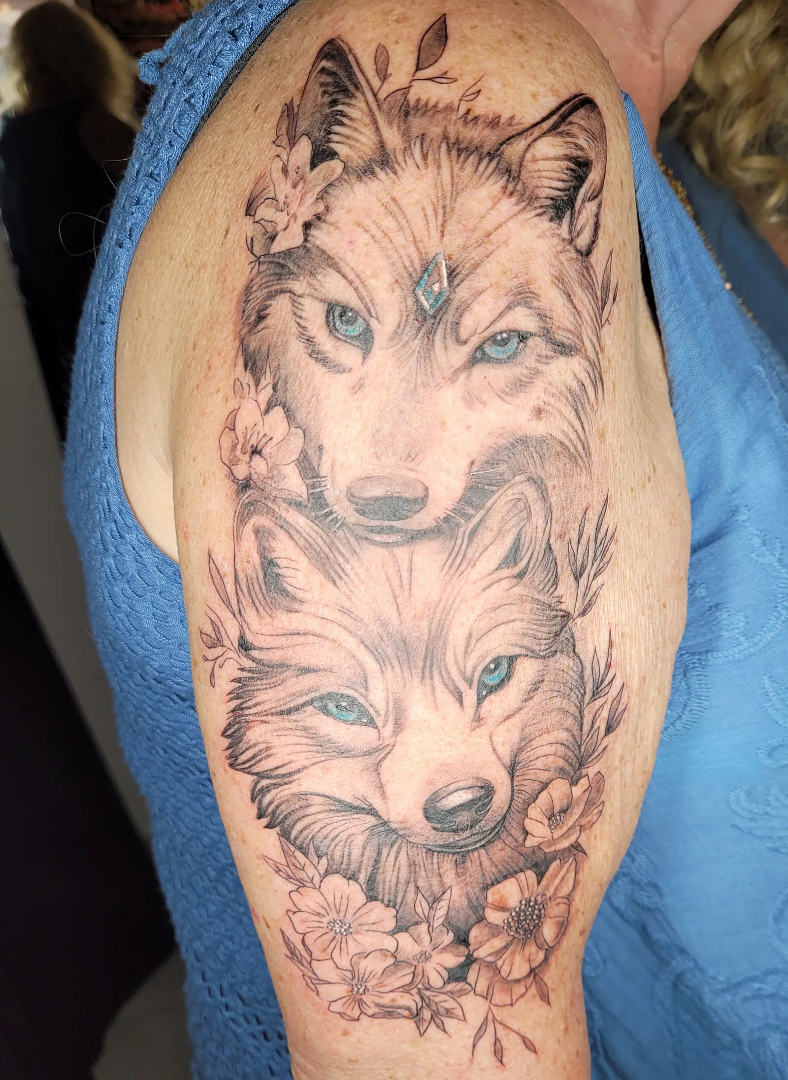 Tatouage réaliste de deux têtes de loups aux yeux bleus entourées de fleurs sur l'épaule, en noir et gris, réalisé par Jean-Marie Joullié au salon JMJ Tattoo proche de Vendres