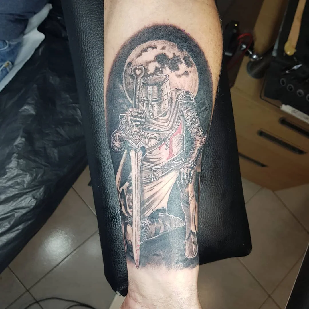 Tatouage réaliste d'un chevalier templier en armure tenant une épée devant une pleine lune, sur l'avant-bras en noir et gris, réalisé par Jean-Marie Joullié au salon JMJ Tattoo à Sérignan