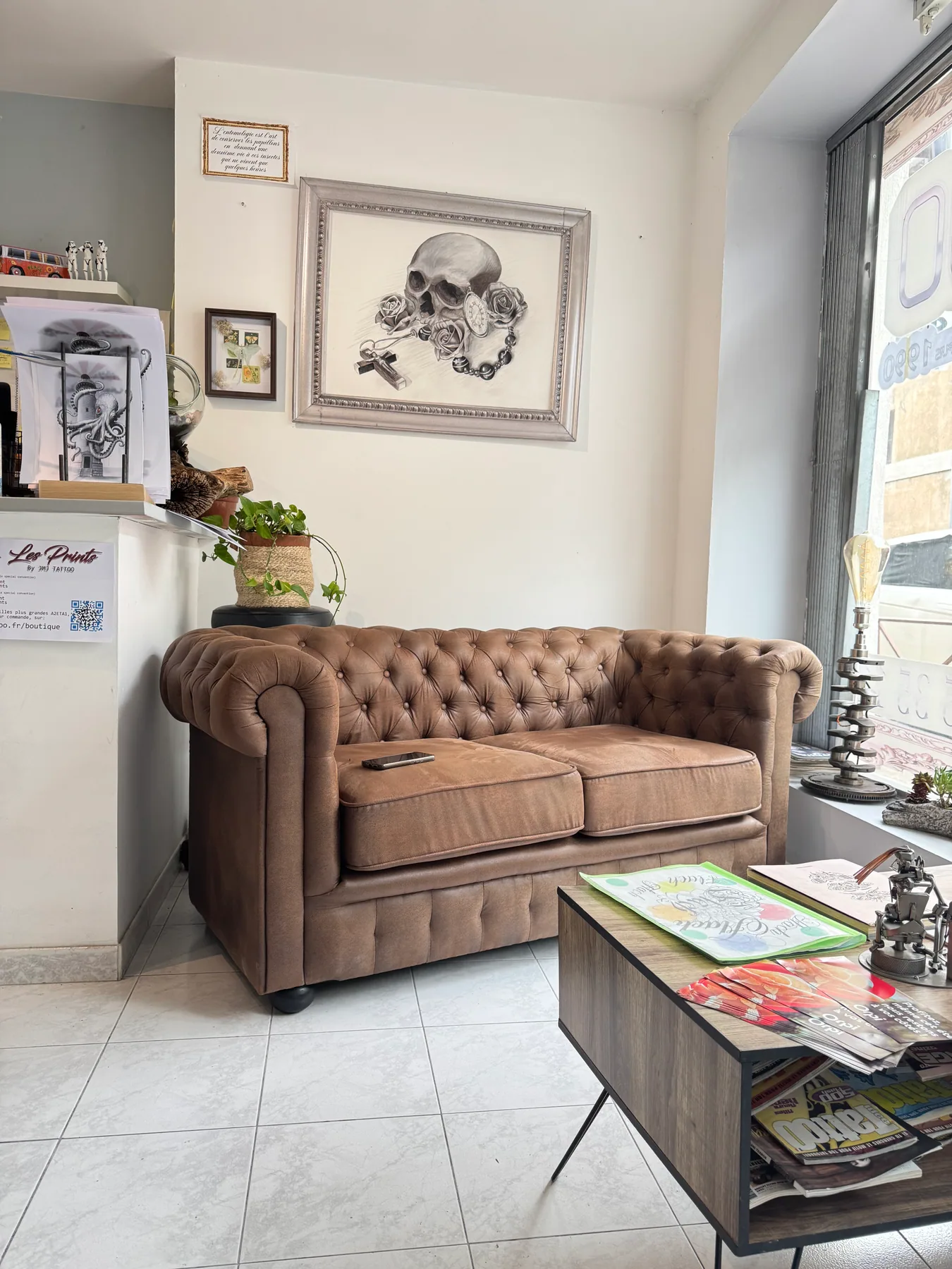 Salle d'attente du salon JMJ Tattoo : canapé Chesterfield cuir et tableau crâne & rose, à l'entrée du salon