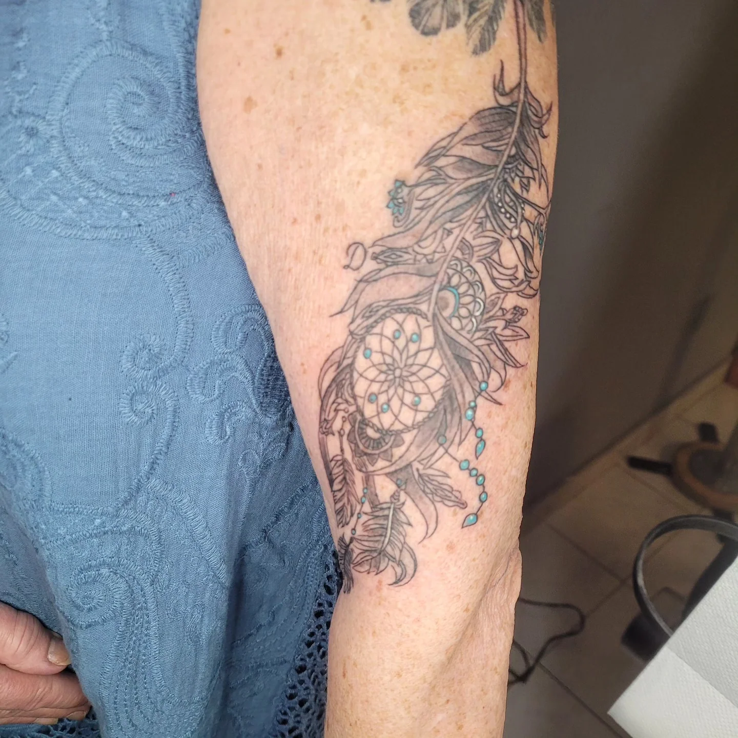 Tatouage d'un attrape-rêve avec plumes et perles turquoise sur le bras, touches de couleur sur noir et gris, réalisé par Jean-Marie Joullié au salon JMJ Tattoo à Béziers et Sérignan
