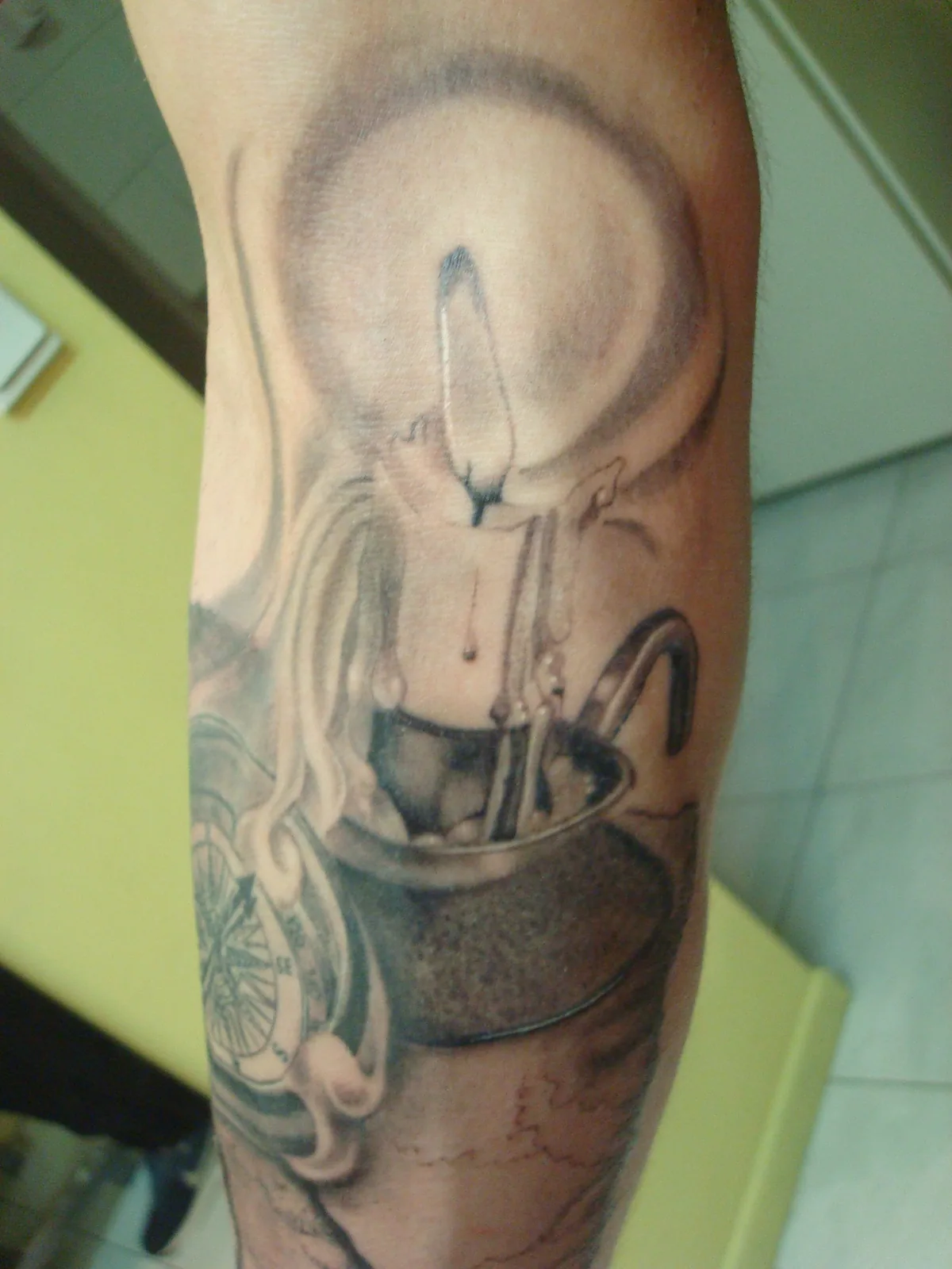 Tatouage sur mesure noir et gris d'une bougie qui se consume au-dessus d'un seau et d'une boussole, composition symbolique sur l'avant-bras, réalisé par Jean-Marie Joullié au salon JMJ Tattoo proche d'Agde