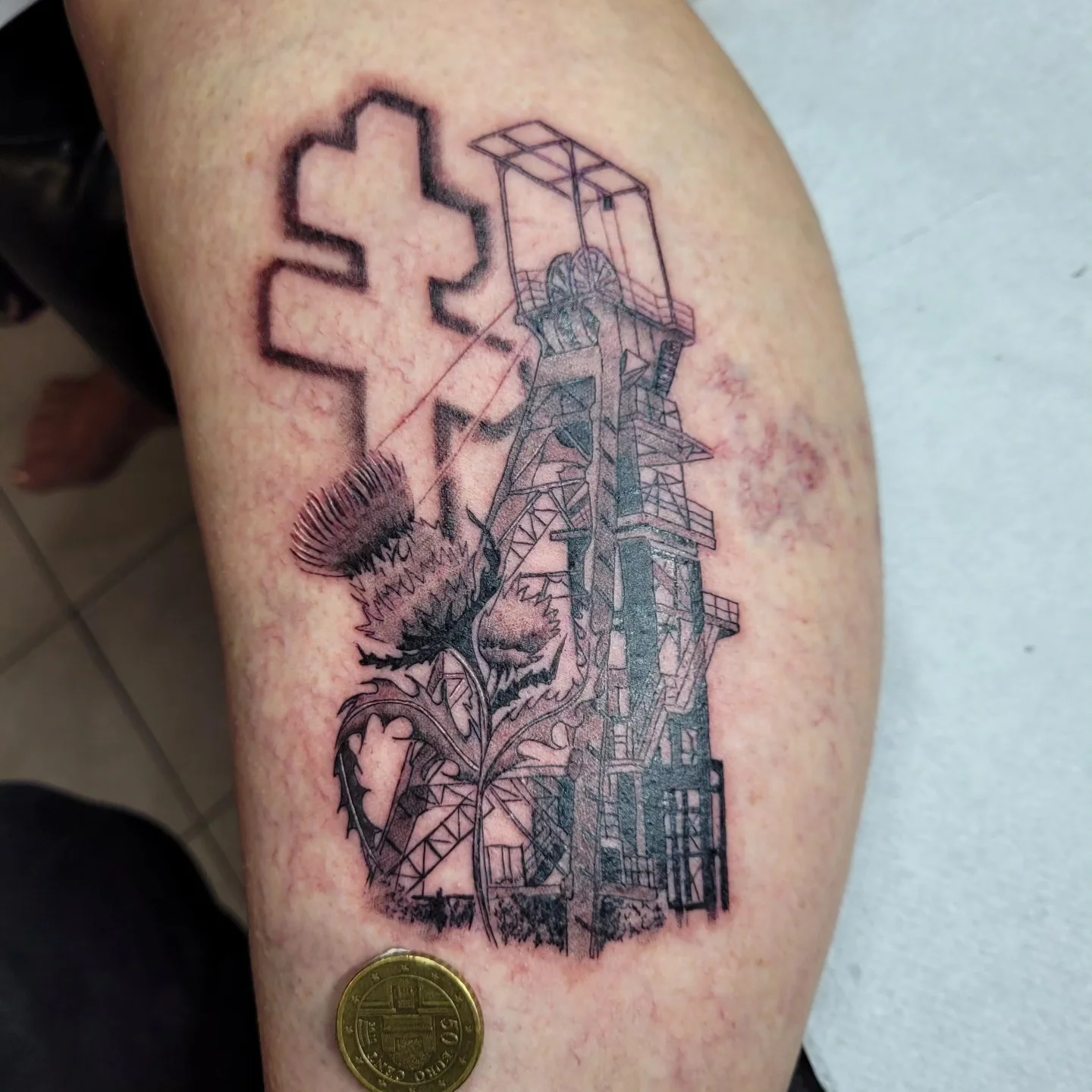 Tatouage hommage au patrimoine minier représentant un chevalement en perspective avec croix et chardon fine line sur le mollet, réalisé par Jean-Marie Joullié au salon JMJ Tattoo à Béziers